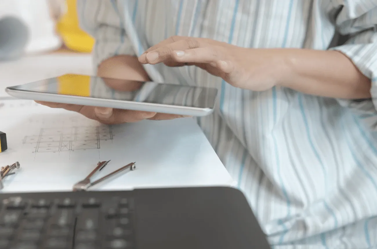 Persona con camisa de rayas usando una tableta sobre planos, con herramientas de dibujo y una laptop en el escritorio