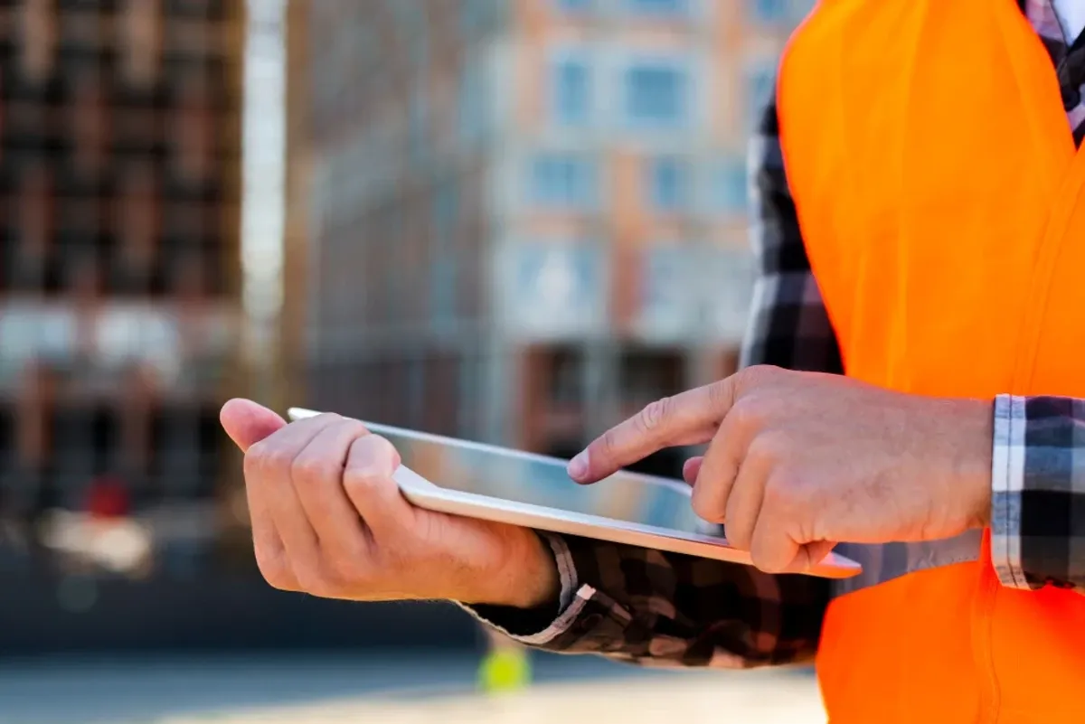 A worker in an orange safety vest and plaid shirt taps a tablet in front of a building under construction