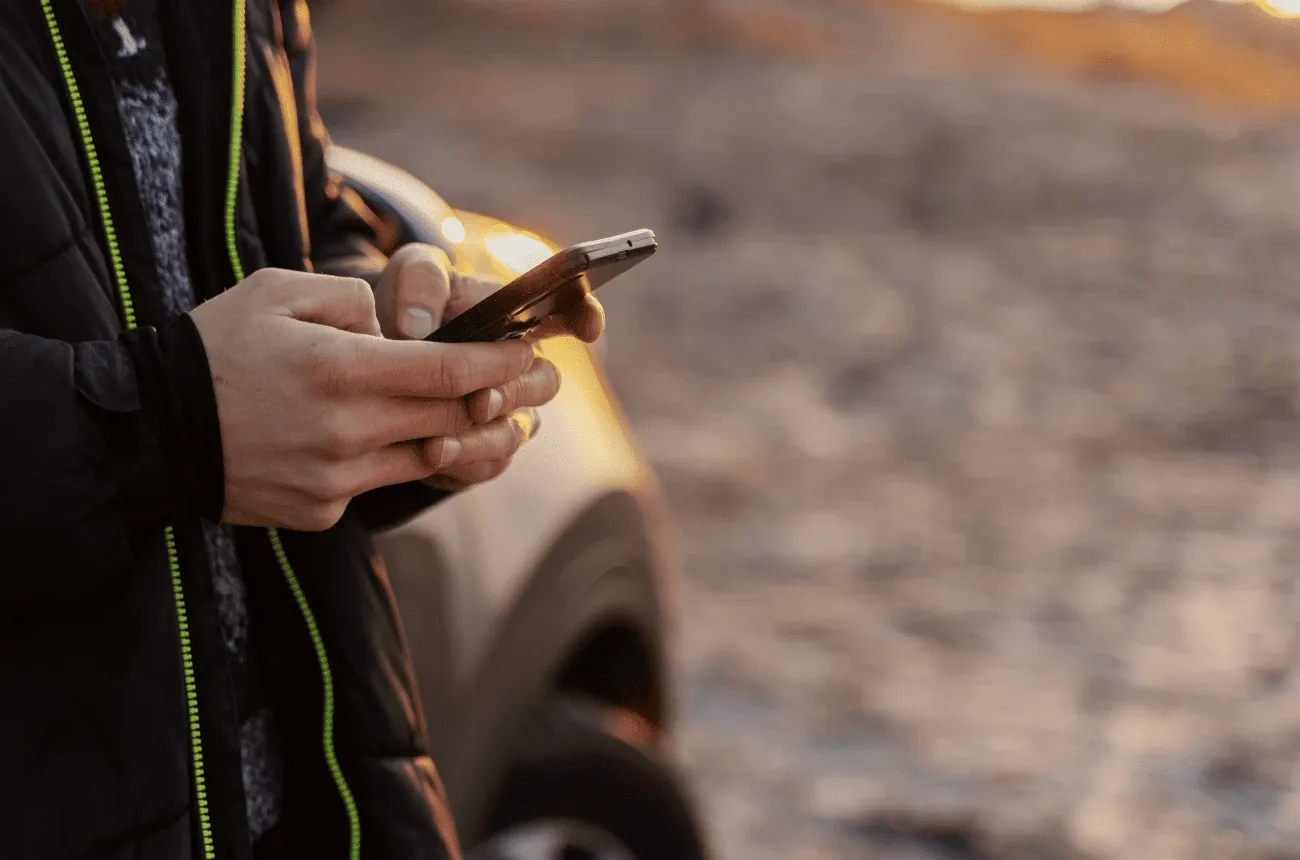 Una persona con chaqueta negra escribe en su teléfono junto a un auto estacionado al atardecer