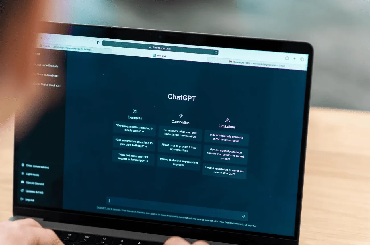 A laptop on a wooden desk showing the ChatGPT start screen, with a person's hands at the keyboard