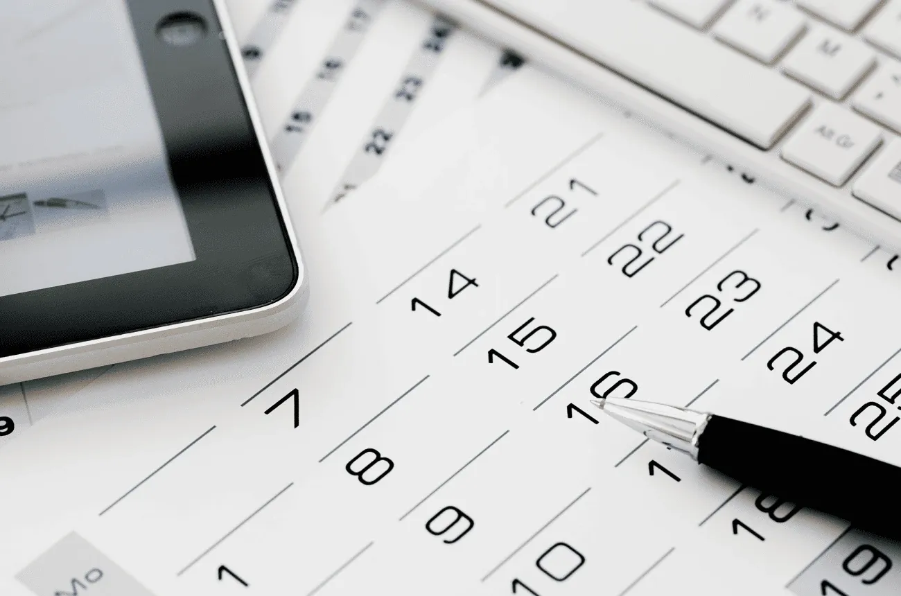 A pen resting on a paper calendar next to a tablet and a white keyboard