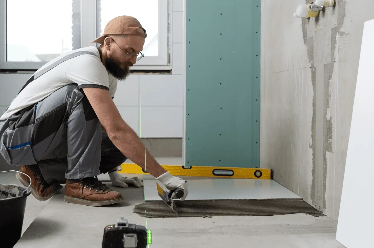 A tiler in a cap and glasses setting a floor tile on notched adhesive in a bathroom under renovation