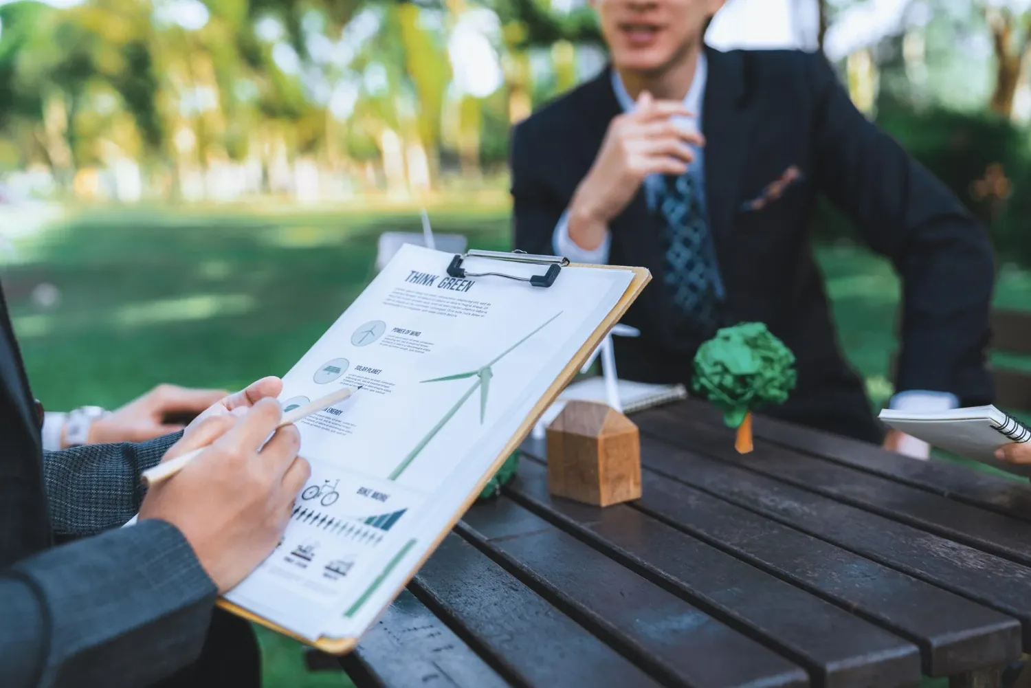Two people in suits at an outdoor table reviewing a green energy handout on a clipboard beside a small wooden house model