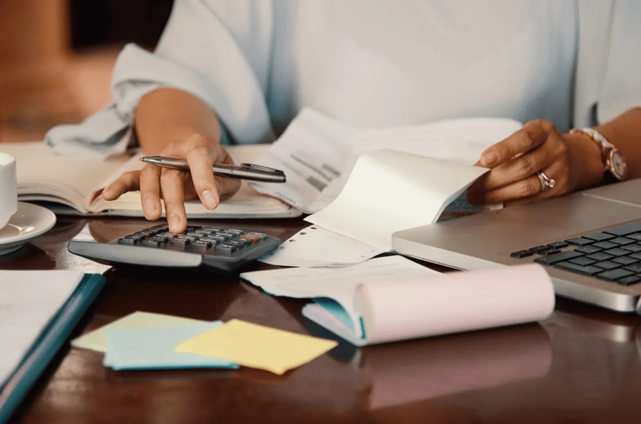 A person working a calculator and holding a pen at a desk covered with receipts, sticky notes and a laptop