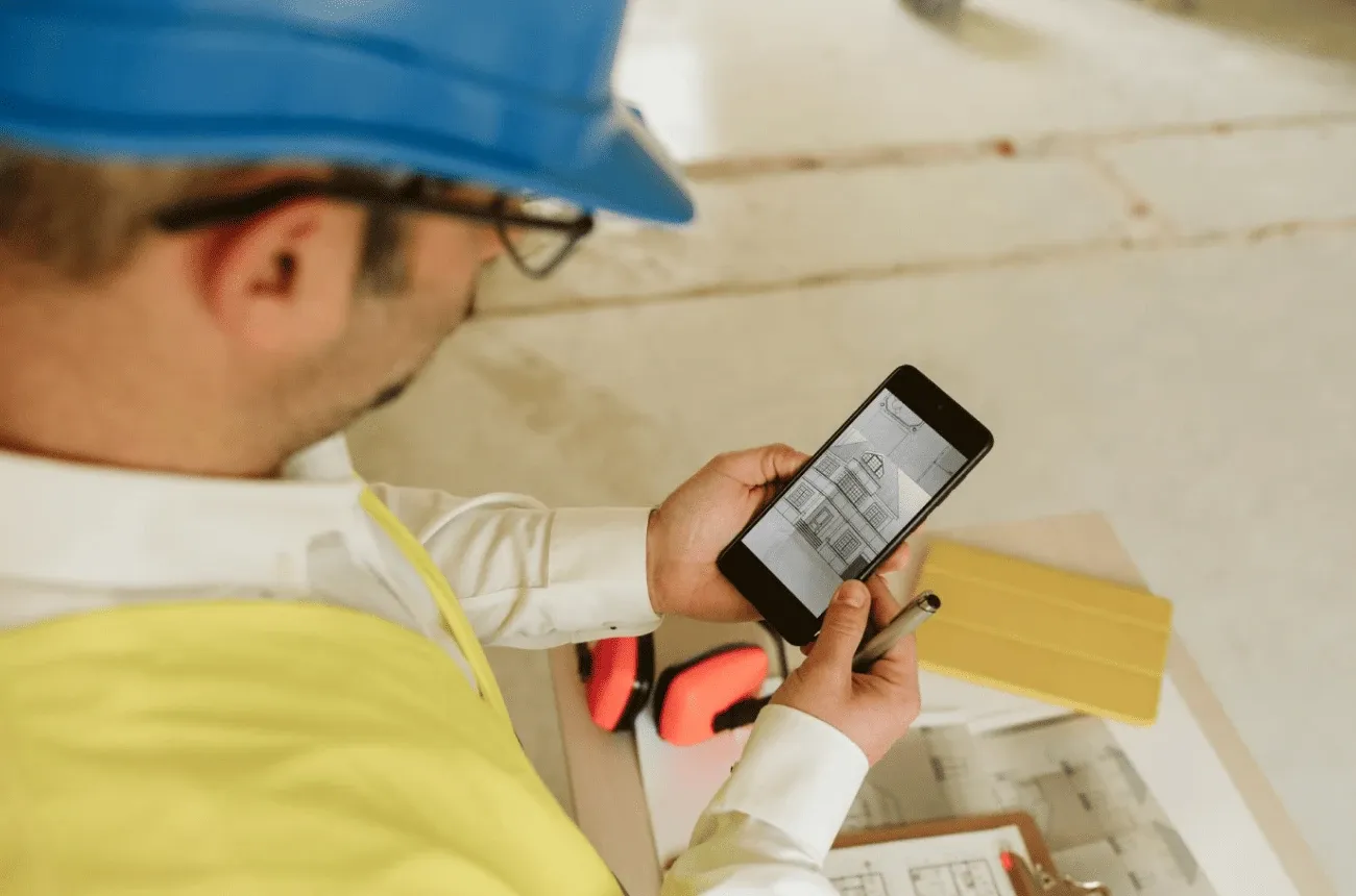 A worker in a blue hard hat and yellow vest looks at a house drawing on his phone above blueprints and ear protectors