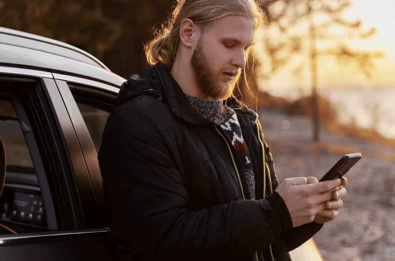A bearded man in a black jacket leans against a car and looks at his phone at sunset