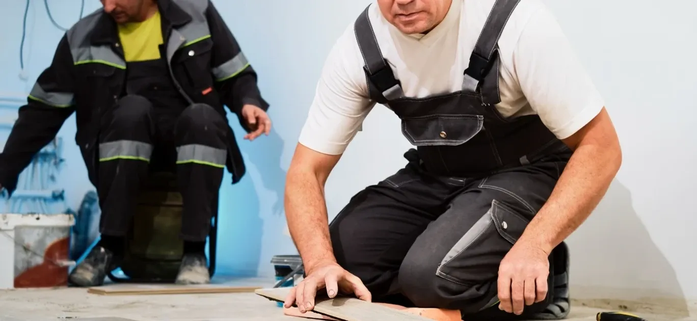 Un trabajador con overol negro se arrodilla para colocar una tabla de piso; detrás, un compañero con ropa reflectante