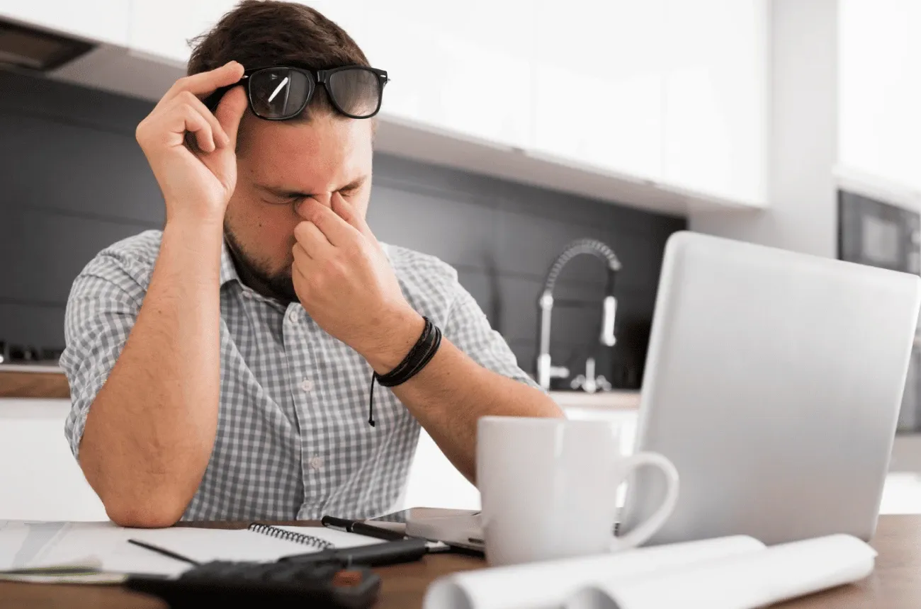 A tired man rubbing his eyes with his glasses pushed up, sitting at a laptop with a mug and a calculator