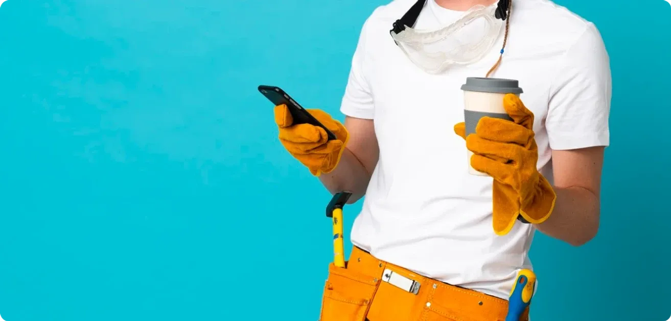 Un trabajador con guantes de cuero, gafas de seguridad al cuello y cinturón de herramientas sostiene un teléfono y un café
