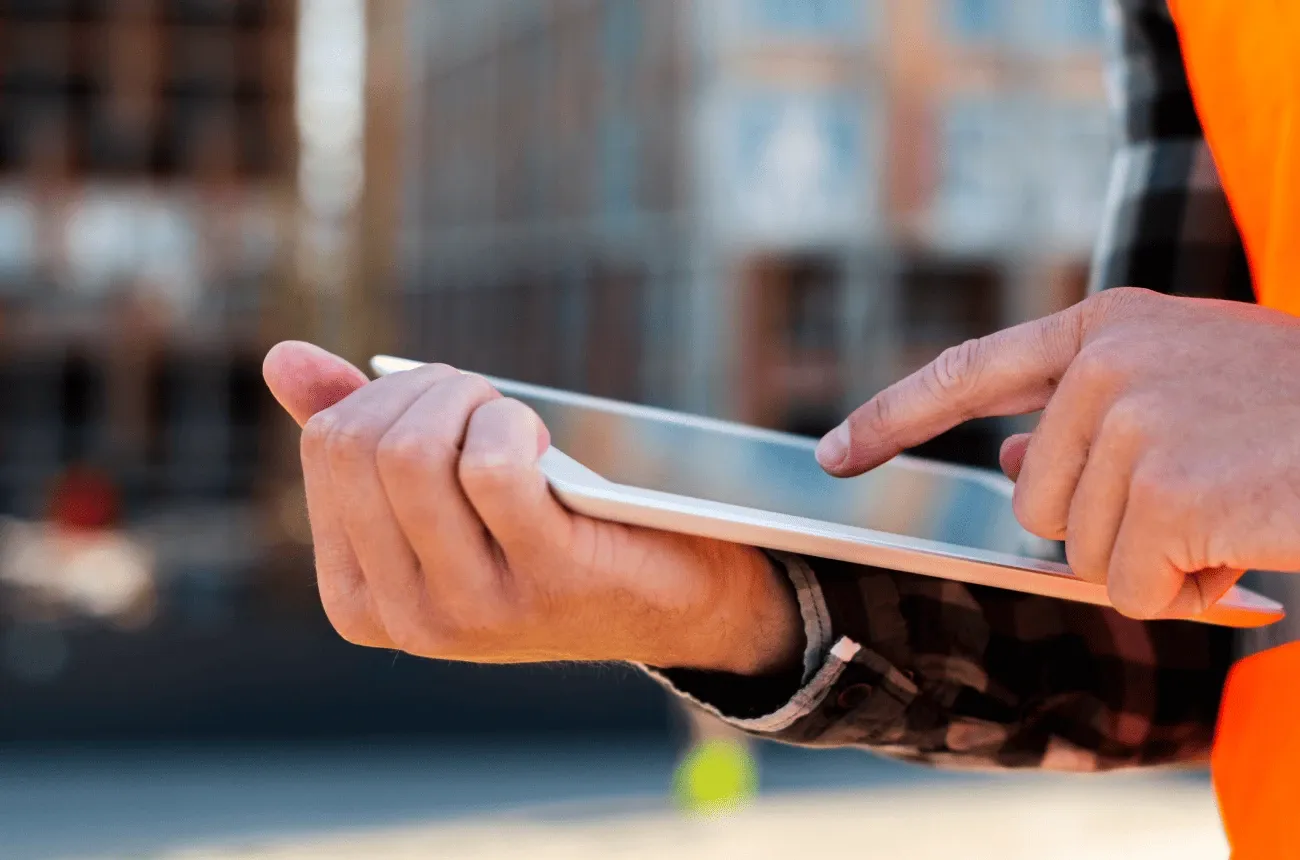 Close-up of a worker in an orange safety vest tapping the screen of a tablet outside a building site