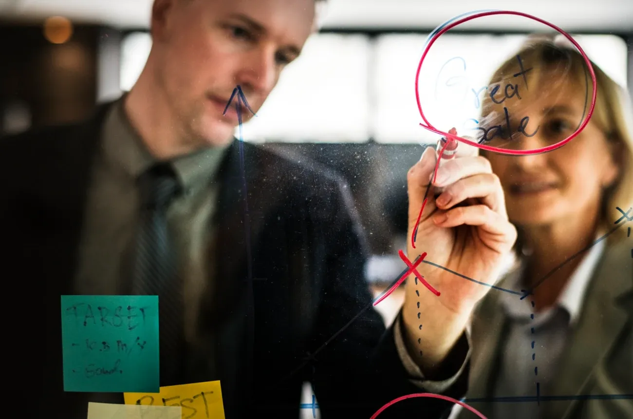 Two colleagues in suits draw a chart on a glass wall with markers, circling the words “Great Sale” in red