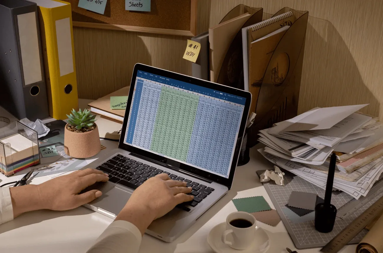 Hands typing on a laptop showing a spreadsheet on a cluttered desk with binders, paper stacks and a cup of coffee