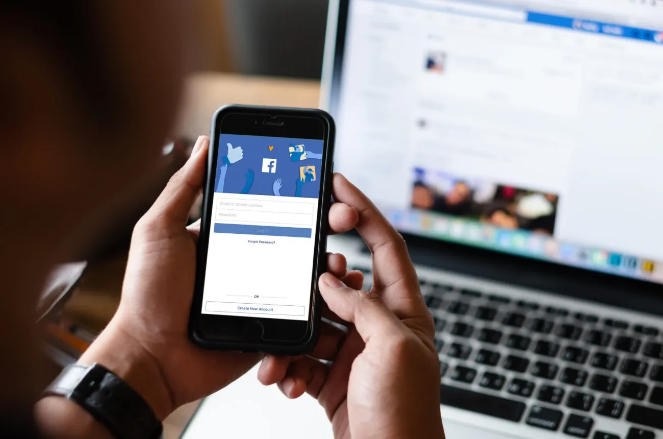 Hands holding a smartphone showing the Facebook login screen in front of an open laptop