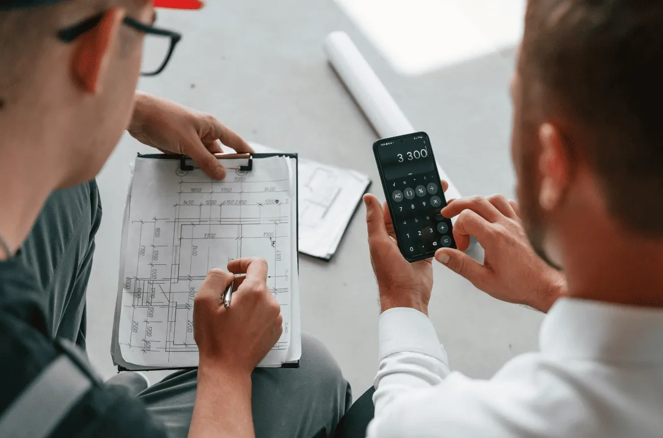 Dos hombres revisan un plano en una tabla con clip mientras uno suma cifras en la calculadora del teléfono