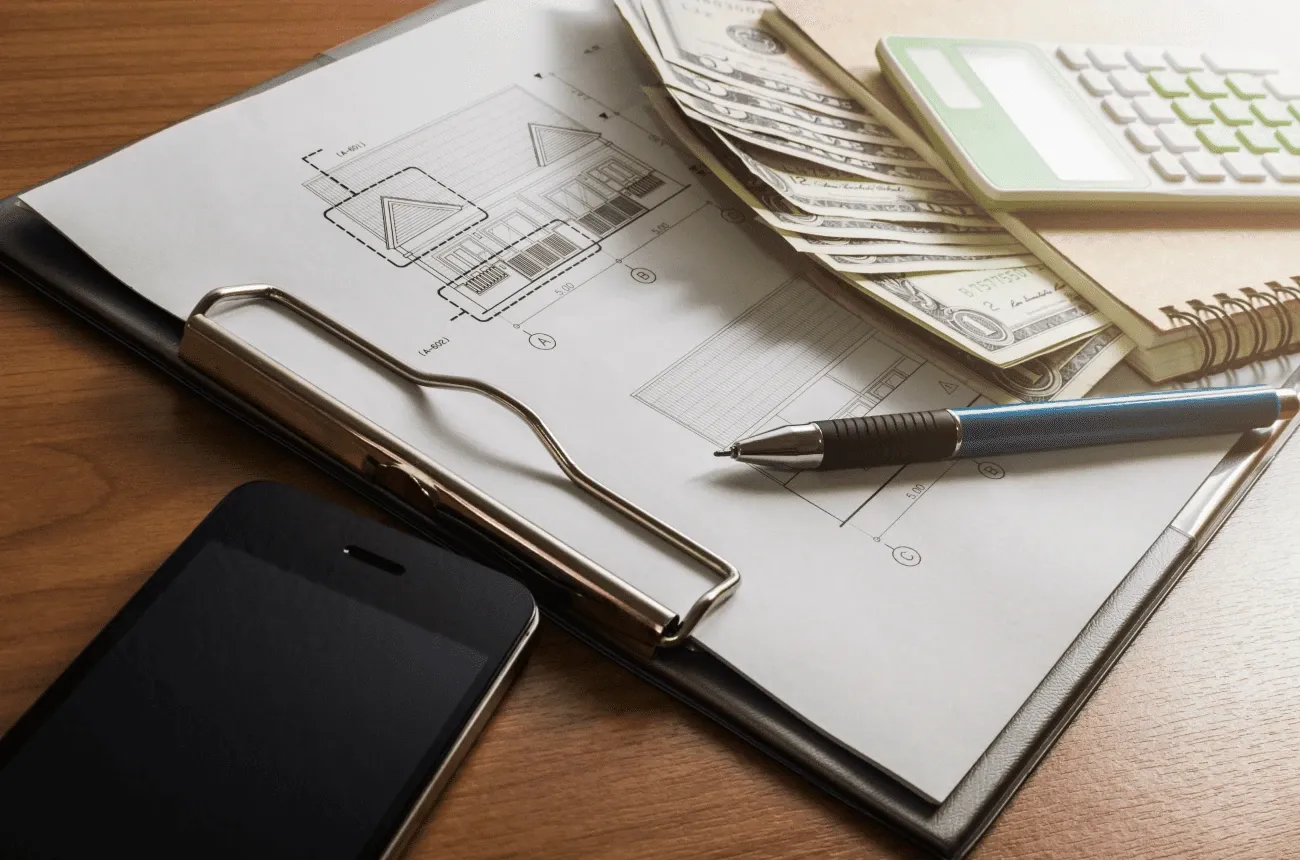 Dibujo de una casa en un portapapeles con bolígrafo, billetes, calculadora, libreta y teléfono sobre una mesa de madera
