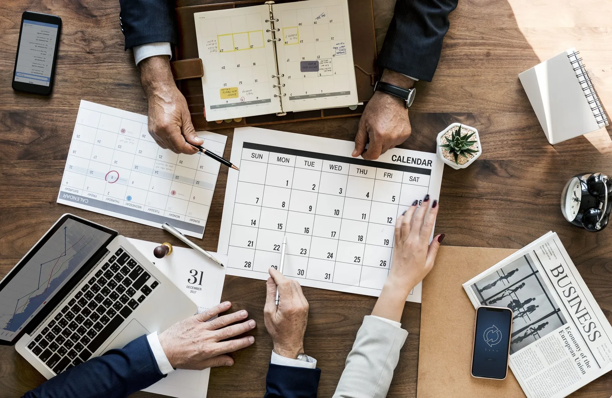 Overhead view of people at a wooden table pointing at paper calendars and a planner next to a laptop and phones