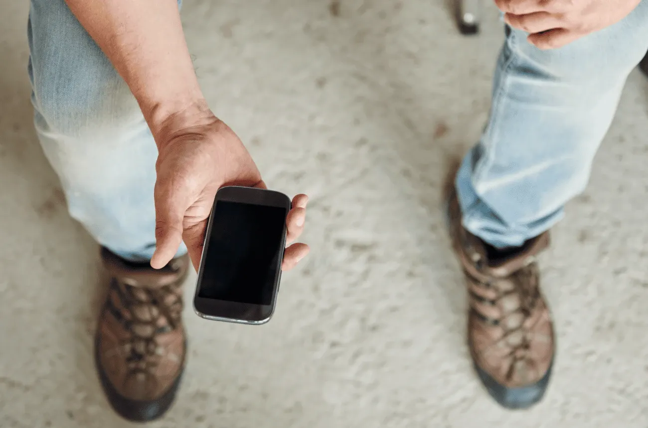 Vista desde arriba de una persona con jeans y botas de trabajo que sostiene un teléfono con la pantalla apagada