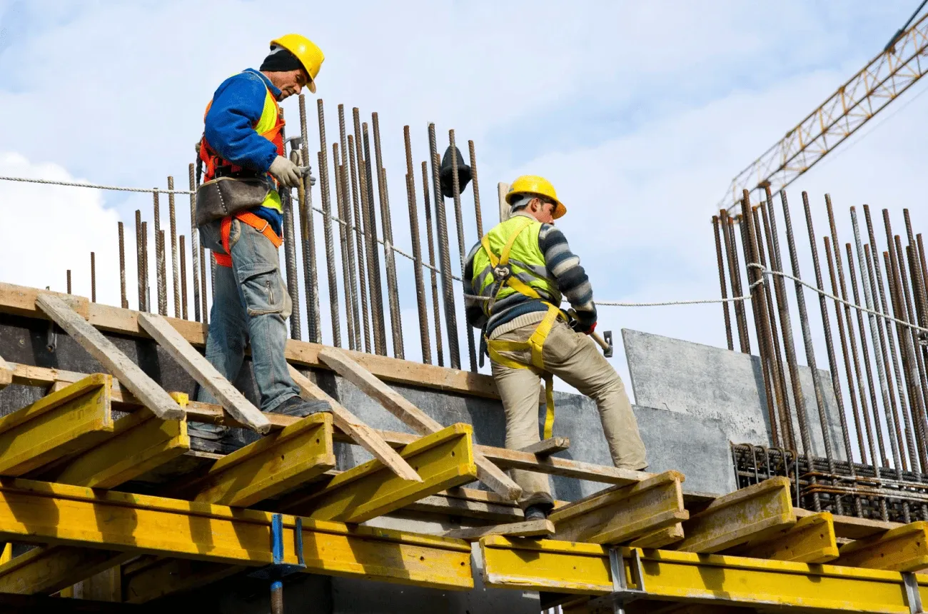 Two construction workers in hard hats and safety vests on formwork among rebar at a building site, with a crane behind