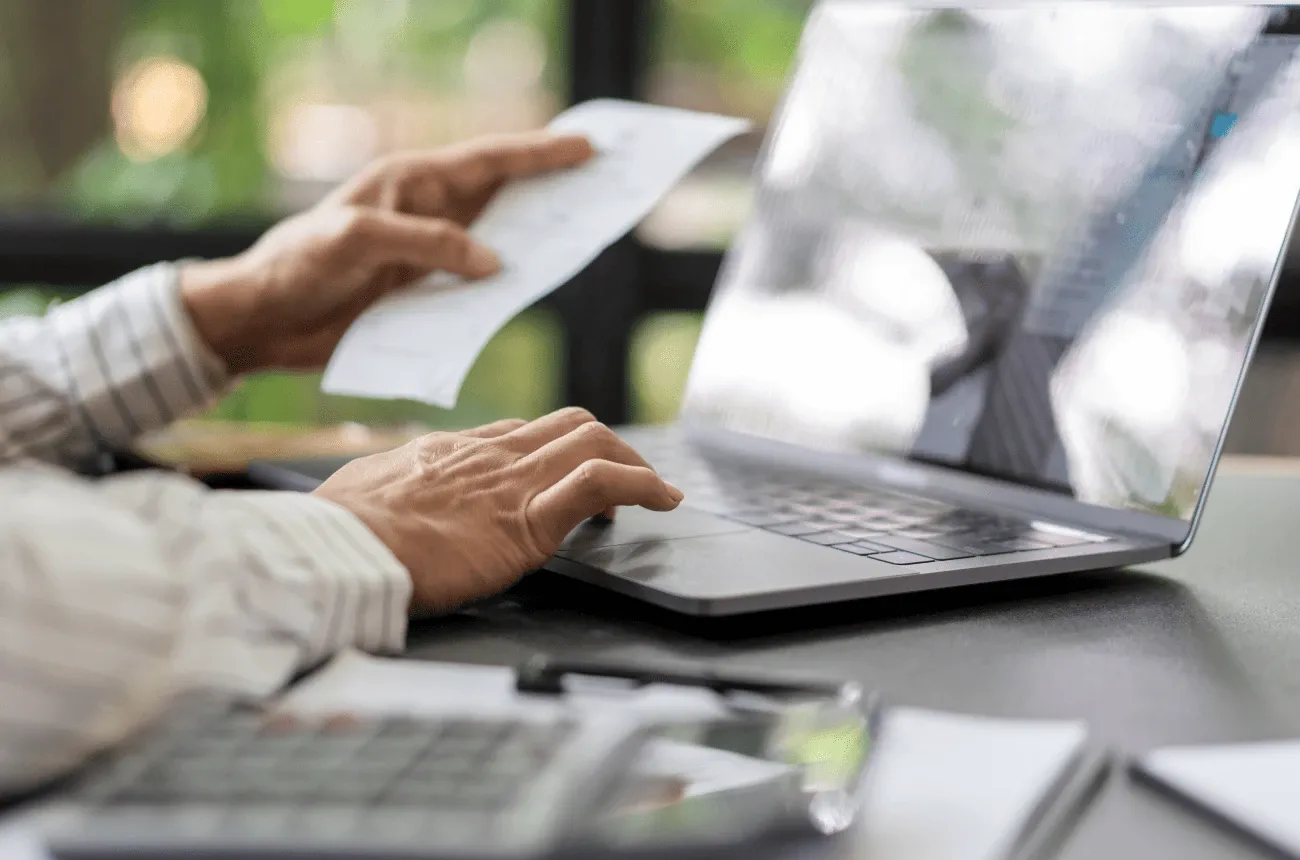 A person holding a paper receipt while using the touchpad of a laptop on a desk with a calculator