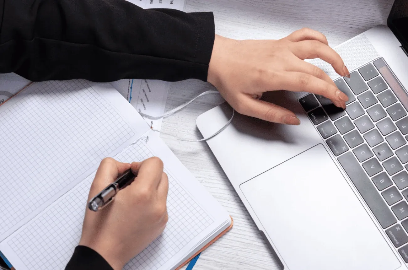 One hand writes in a grid notebook while the other types on a laptop keyboard on a white desk