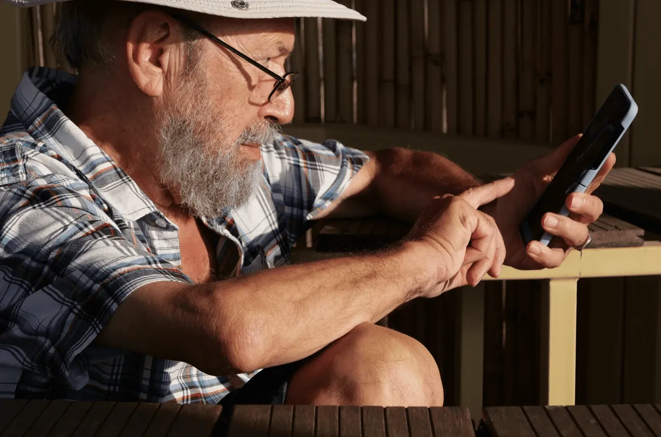An older bearded man in a hat, glasses and a plaid shirt tapping on a smartphone while sitting outdoors in the sun