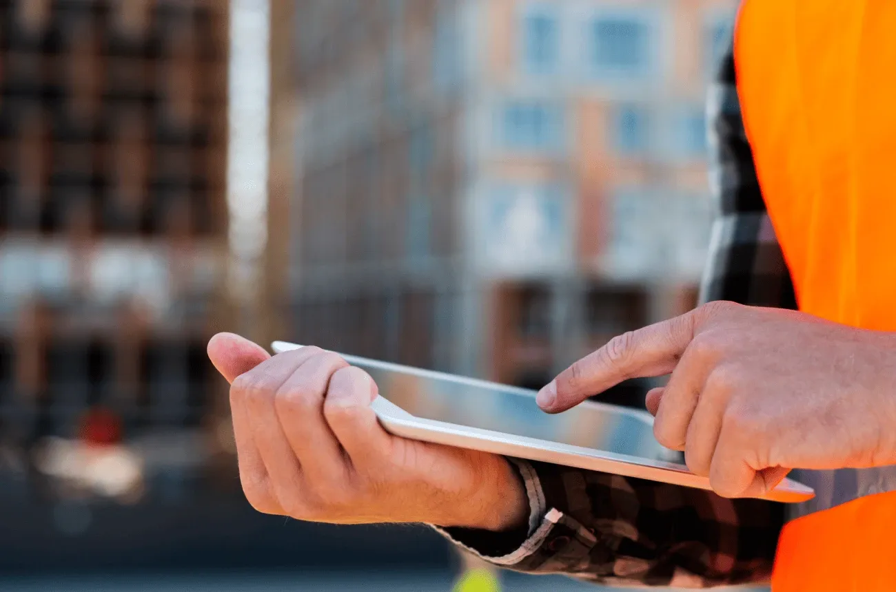 A worker in an orange safety vest taps on a tablet in front of a blurred building under construction