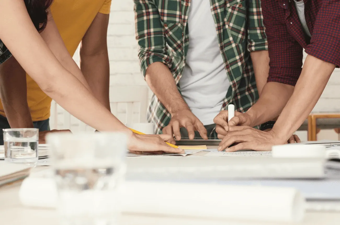 Several people in casual shirts lean over a table and mark up plans with a pencil and a marker