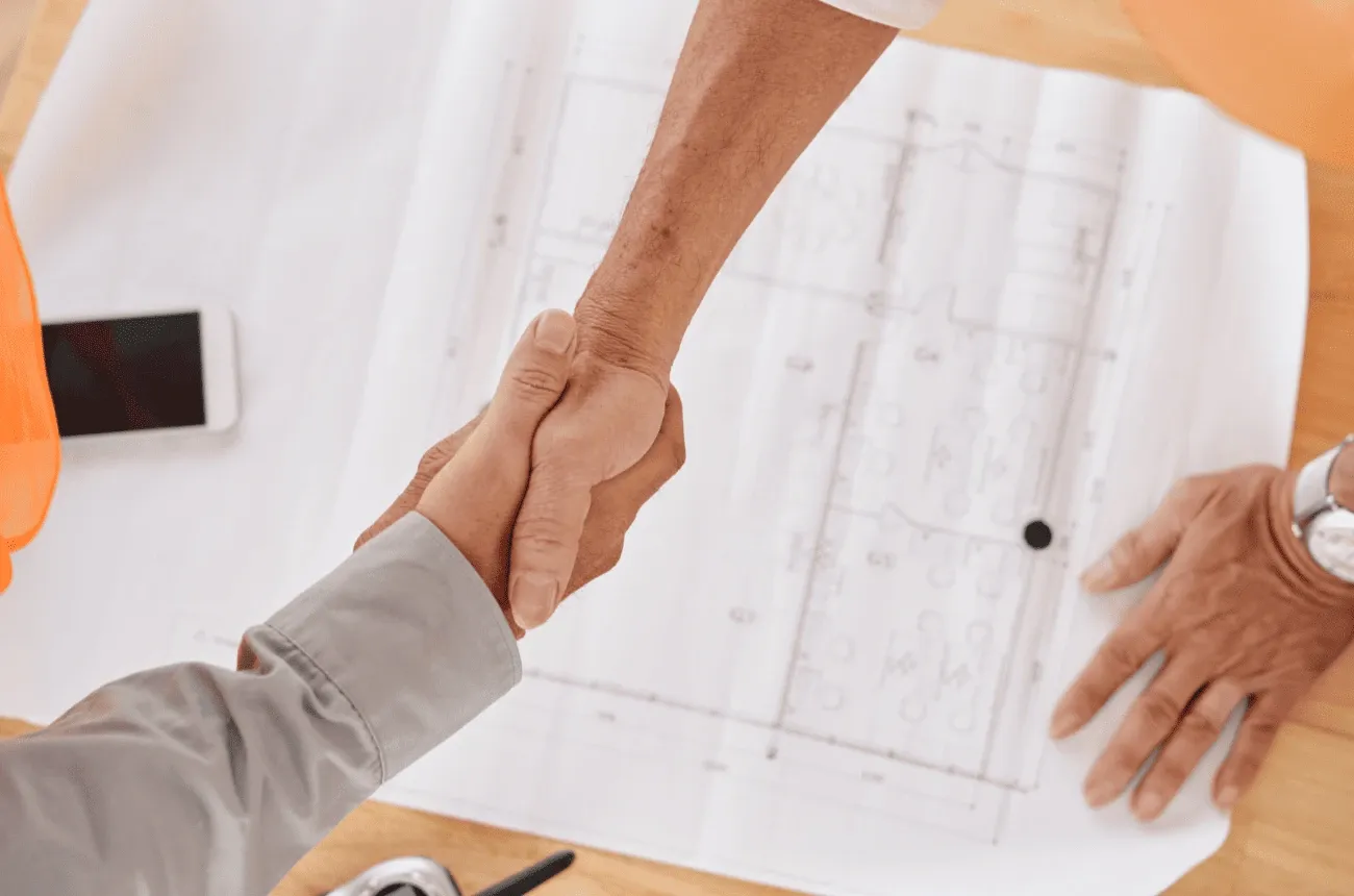 Dos personas se dan la mano sobre un plano de construcción en una mesa junto a un teléfono