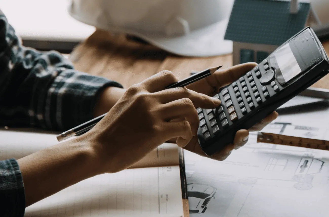 Hands holding a pencil and a calculator over house plans, with a hard hat, a tape measure and a model house nearby