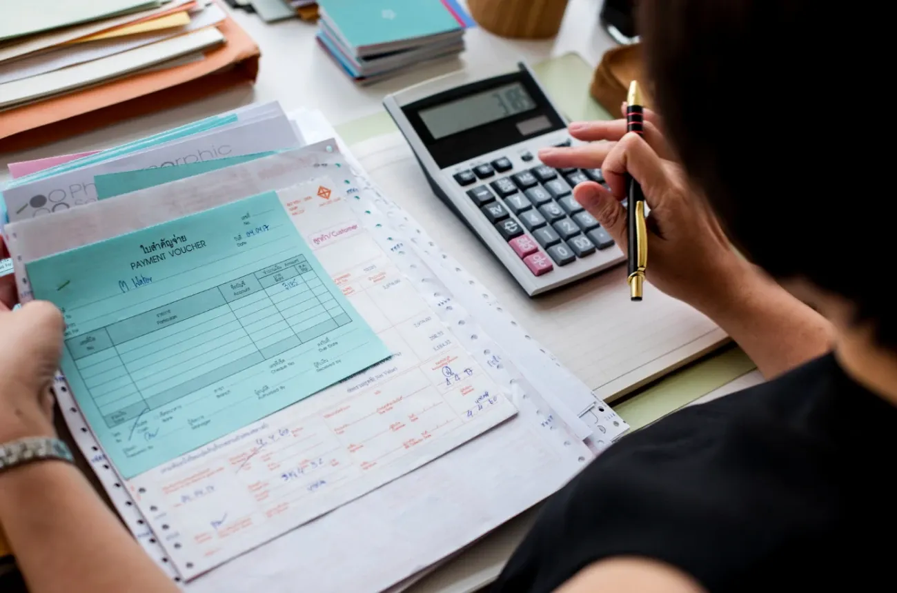 A person with a pen using a calculator over a stack of payment vouchers and invoices on a desk