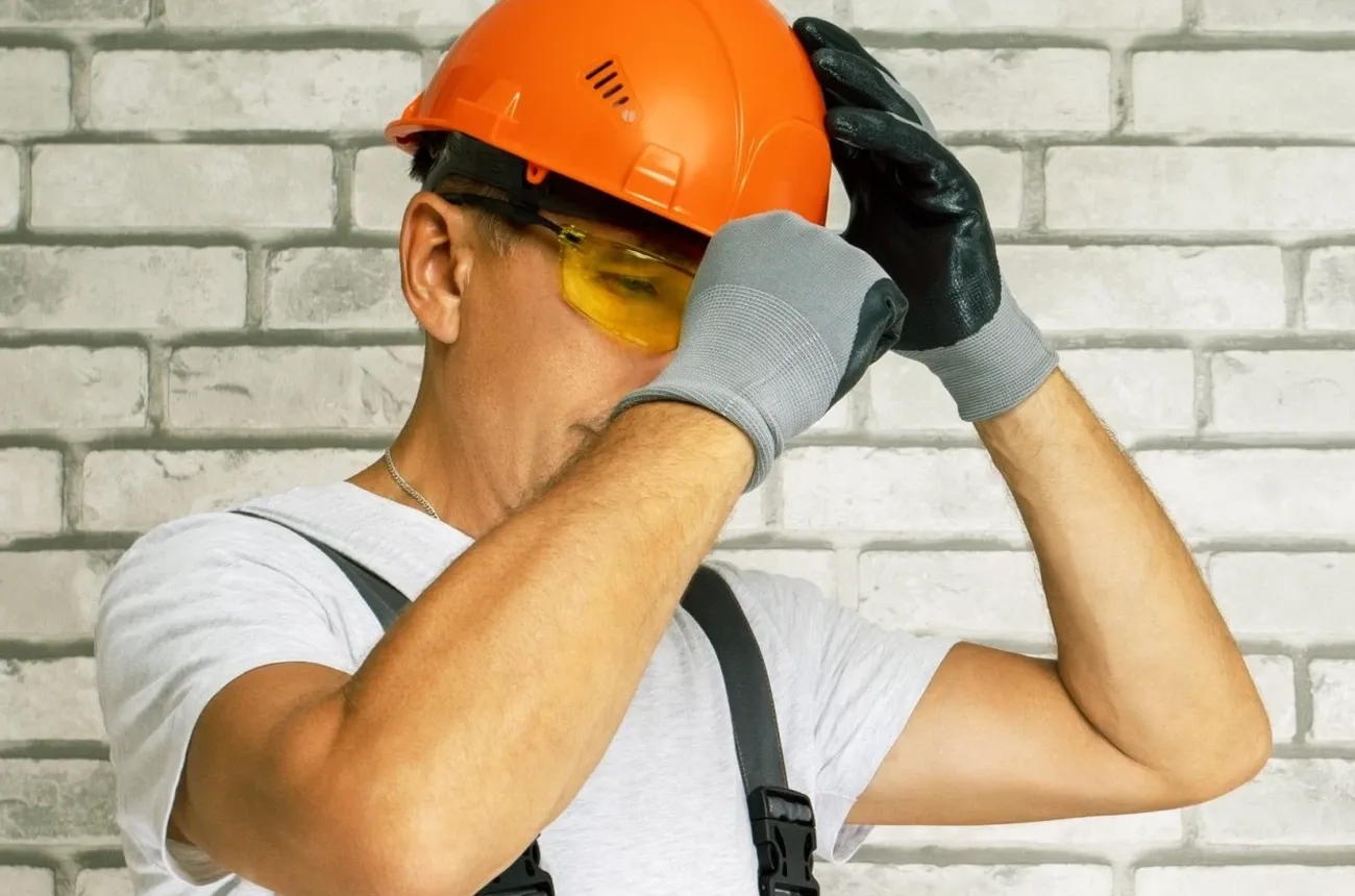 A worker in a white T-shirt and overalls adjusts an orange hard hat, wearing yellow safety glasses and work gloves