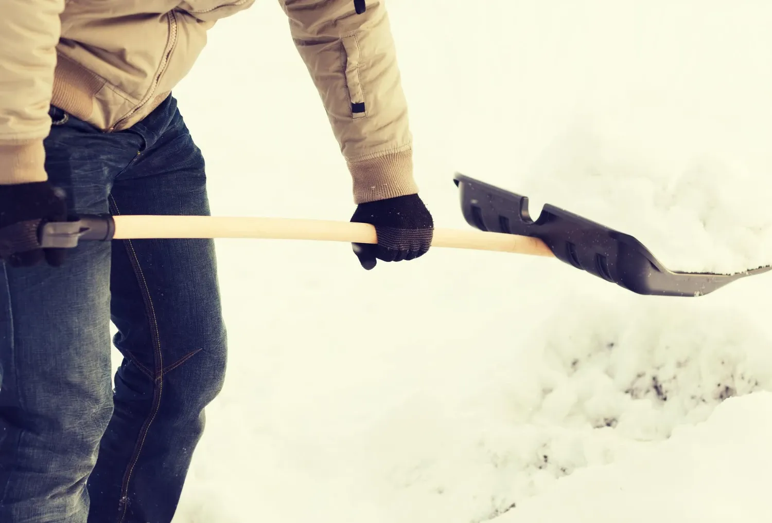 Una persona con chaqueta beige, jeans y guantes negros retira nieve con una pala de mango de madera