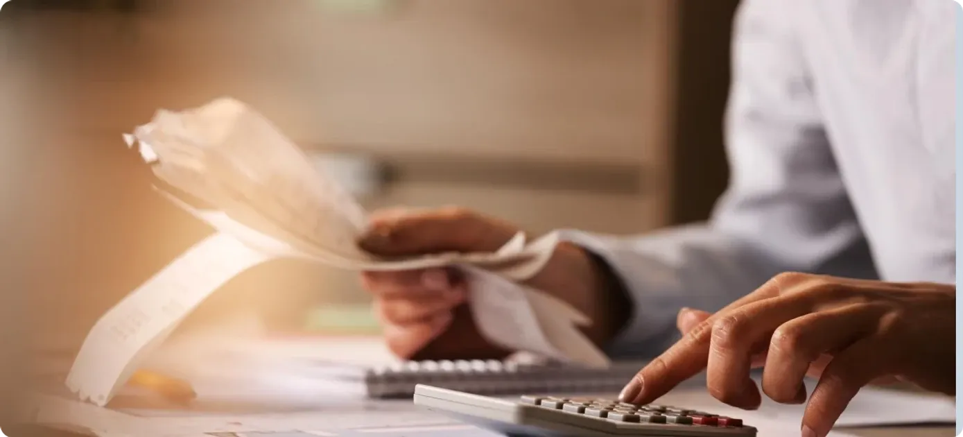 A person holding a stack of receipts and typing on a calculator at a desk