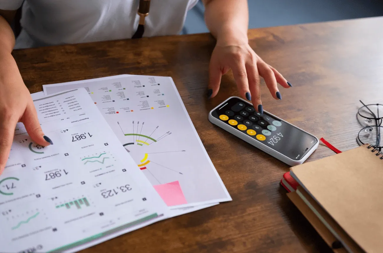 A woman taps a phone calculator on a wooden desk next to printed reports with charts and a notebook