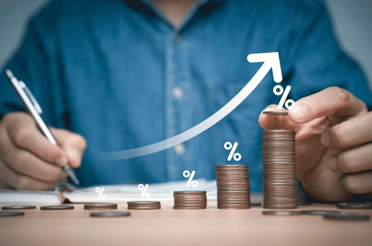 A hand stacking coins in rising piles marked with percent signs under an upward arrow, while another hand writes notes