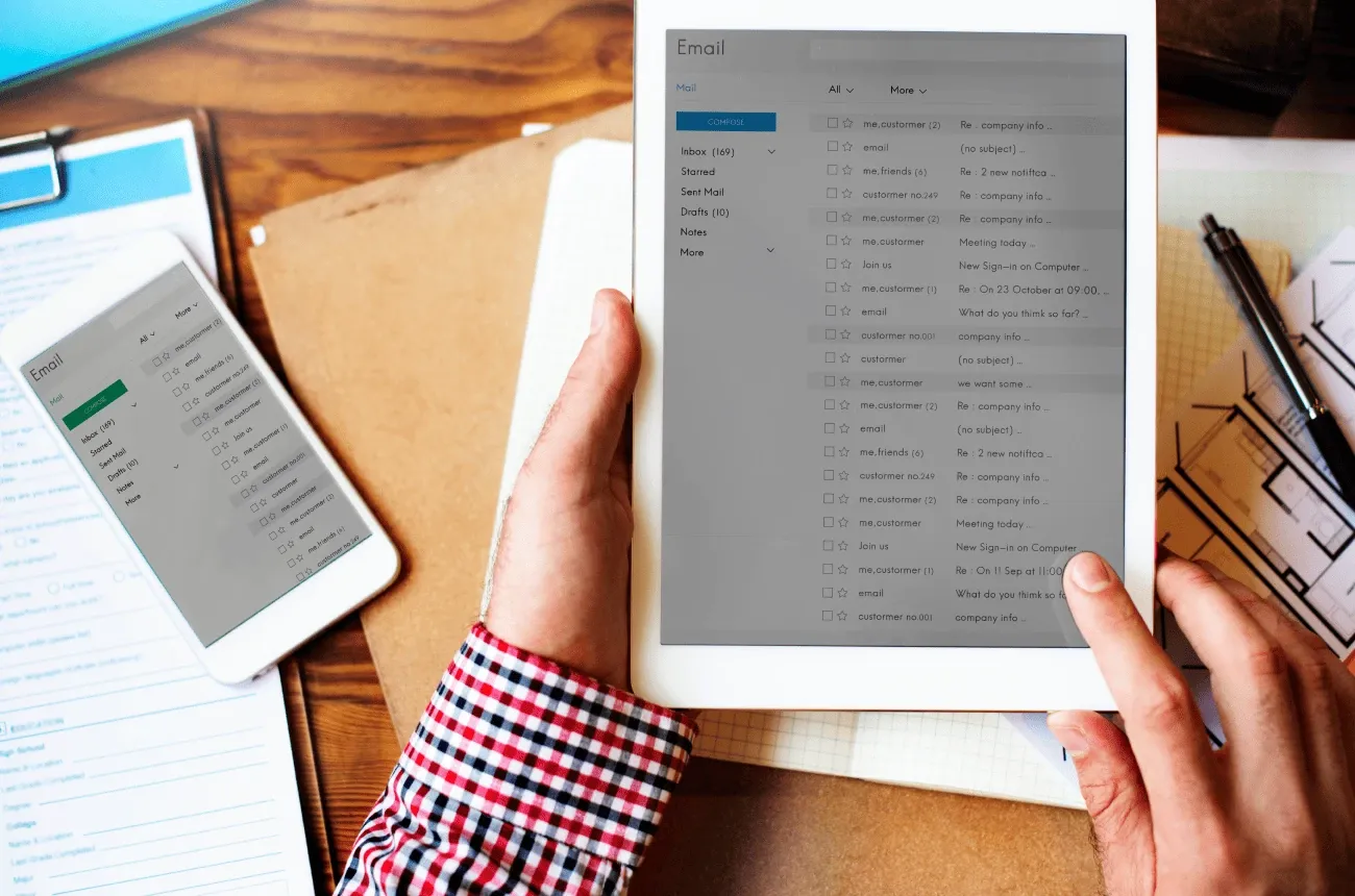 Hands holding a tablet with an email inbox open above a desk with a phone, forms and a floor plan
