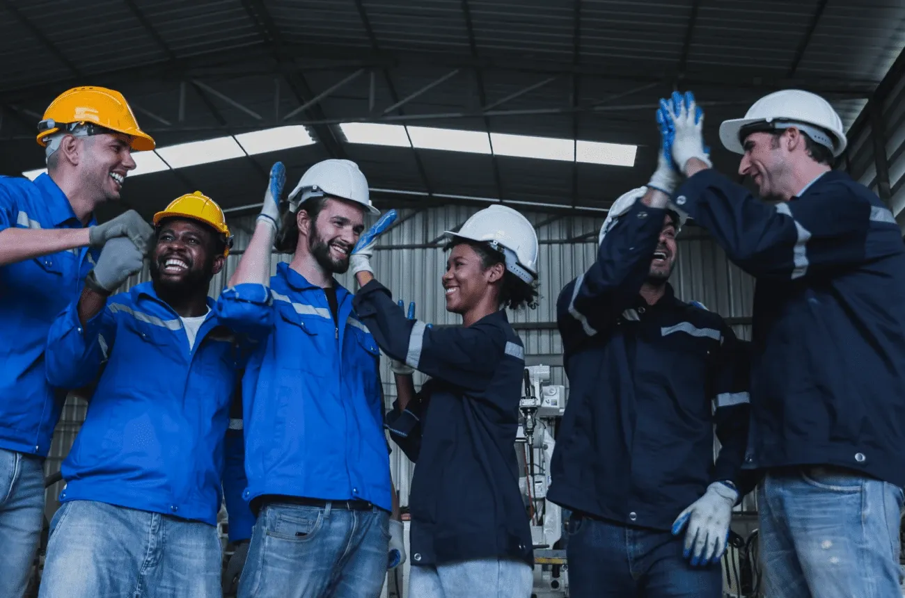 Smiling workers in hard hats and blue work jackets high-fiving each other inside a warehouse