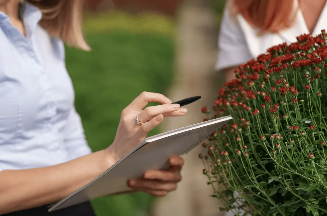 A woman holding a pen and notepad stands next to a pot of red flowers in a garden