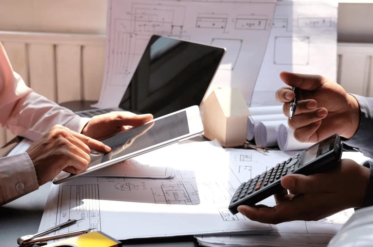 Dos personas revisan planos en un escritorio, una con una tableta y la otra con una calculadora y un bolígrafo