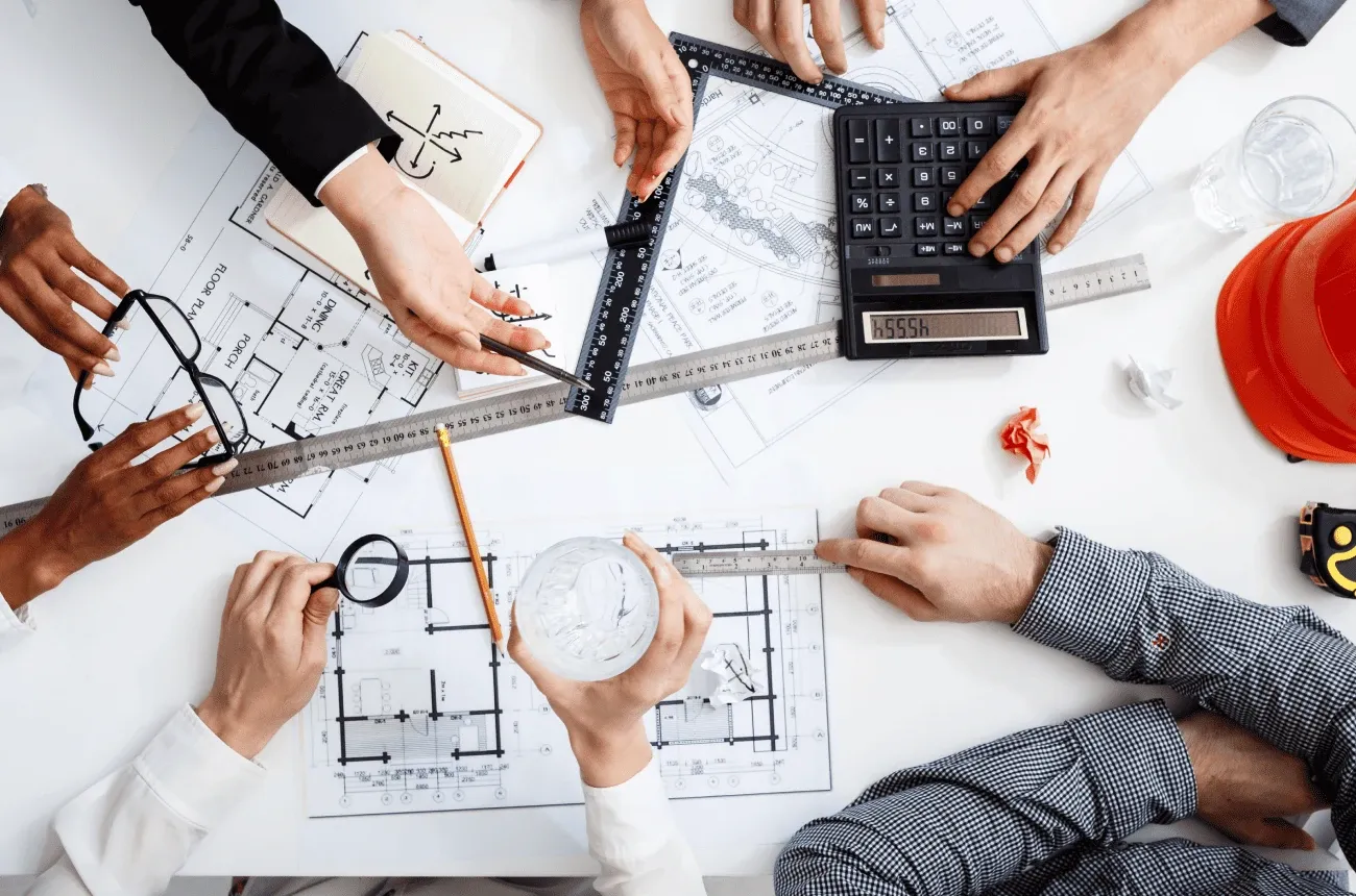 Vista superior de varias manos sobre planos con una calculadora, reglas, una lupa y un casco de obra