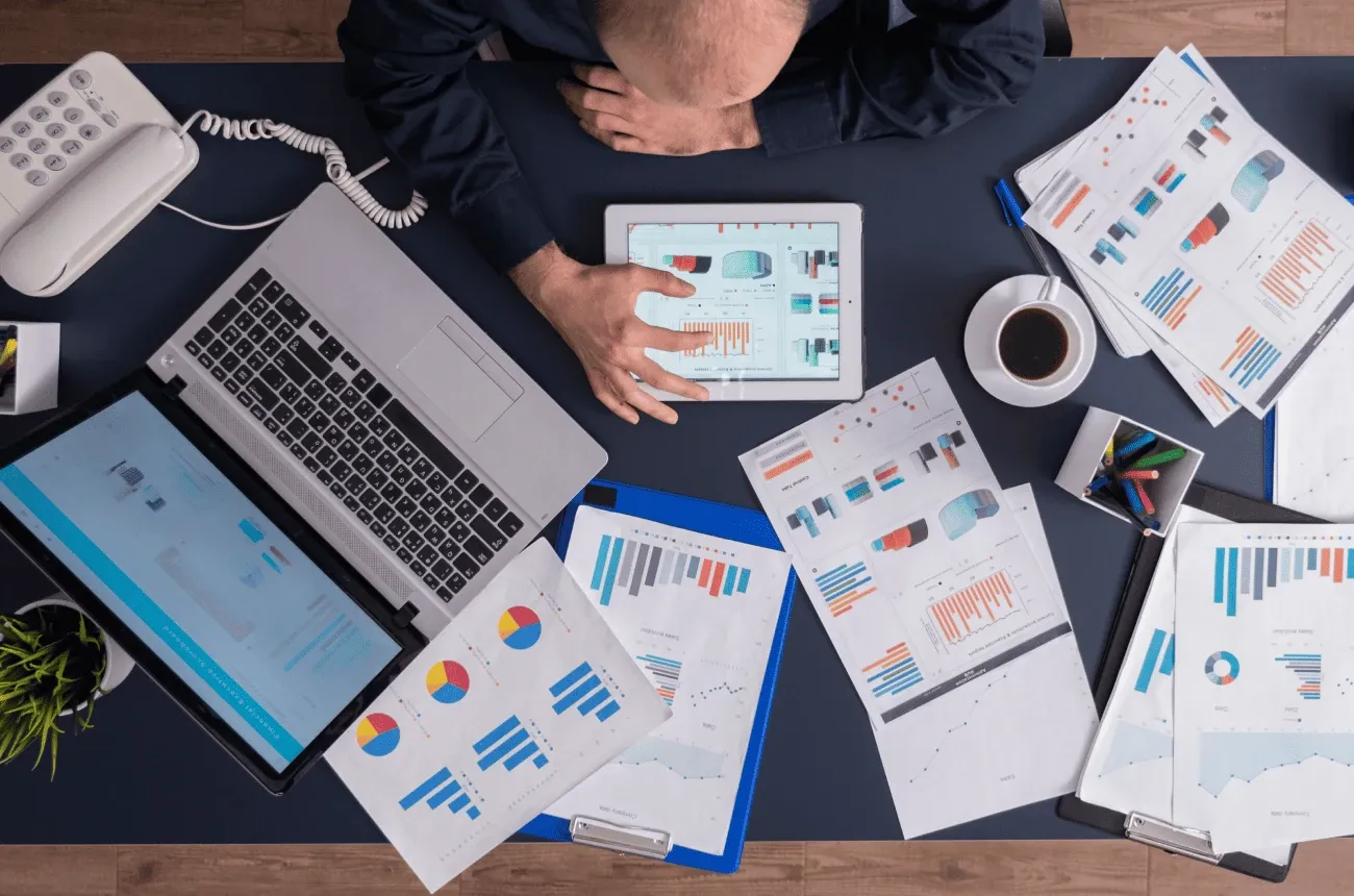 Overhead view of a person at a desk using a tablet, surrounded by printed charts, a laptop, a desk phone and a coffee