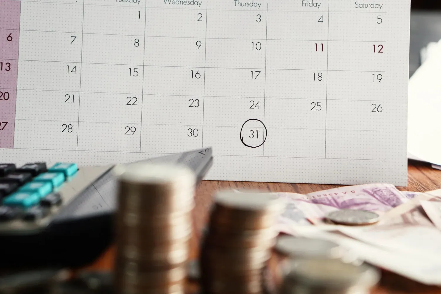 A desk calendar with the 31st circled, behind stacks of coins, banknotes and a calculator