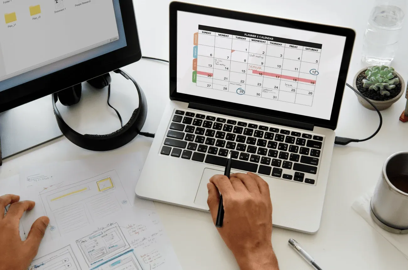 A hand holding a pen next to a laptop showing a monthly planner calendar, with paper sketches, a monitor and a plant