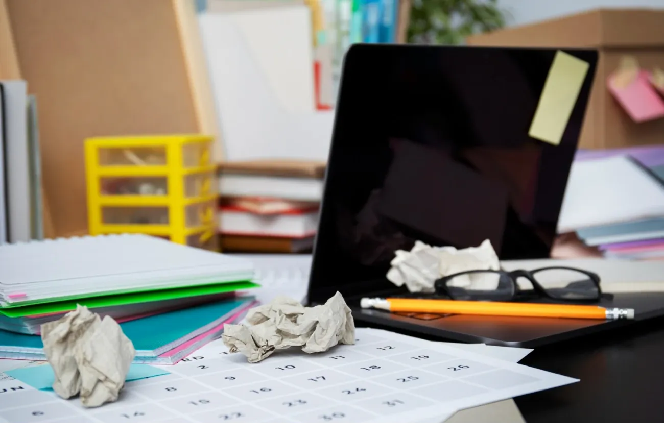 A messy desk with crumpled paper on a calendar, a laptop, glasses, a pencil and stacked notebooks