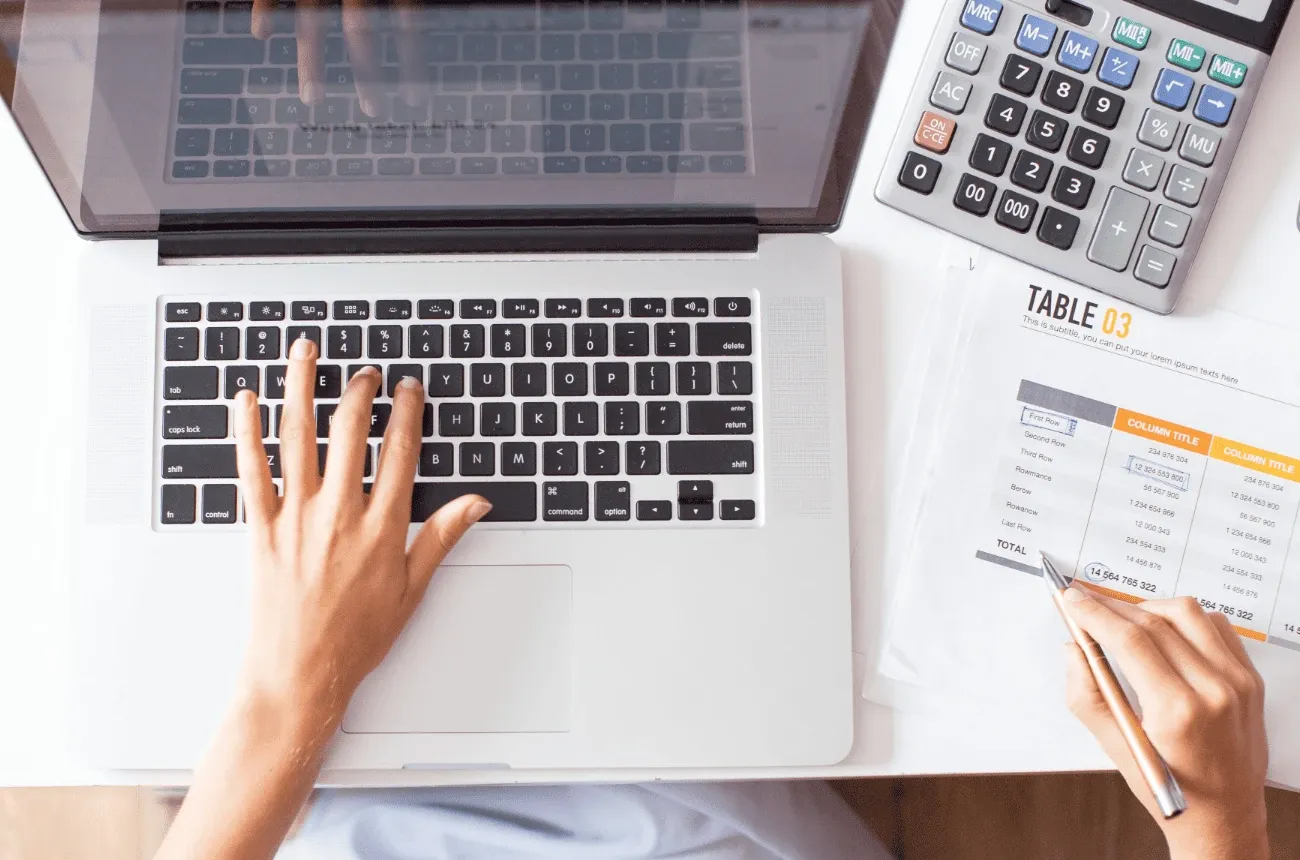 Una mano escribe en una laptop y la otra revisa con un bolígrafo una tabla de cifras impresa junto a una calculadora