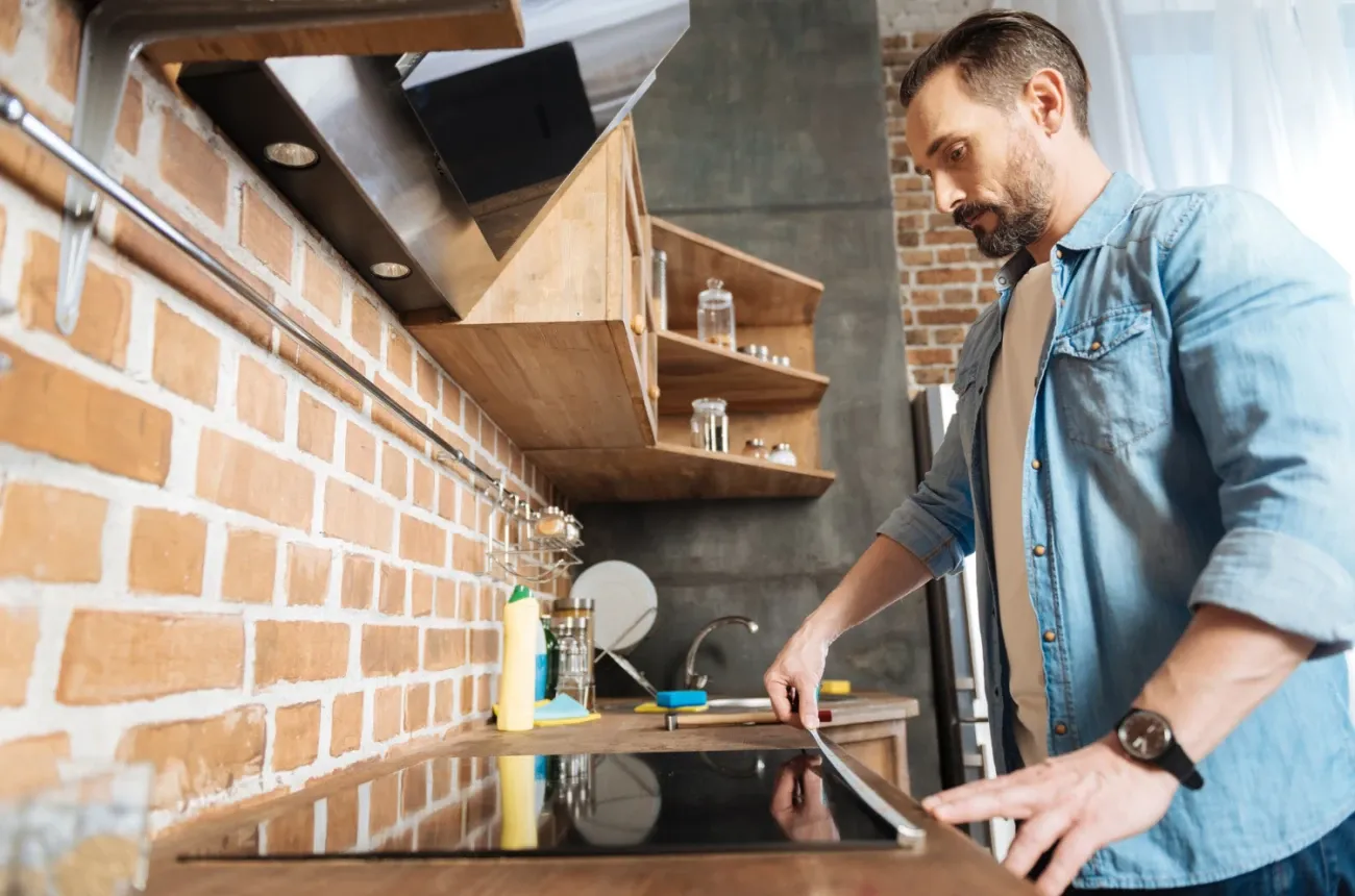 A bearded man in a denim shirt measuring a cooktop on a wooden counter in a kitchen with a brick wall and open shelves