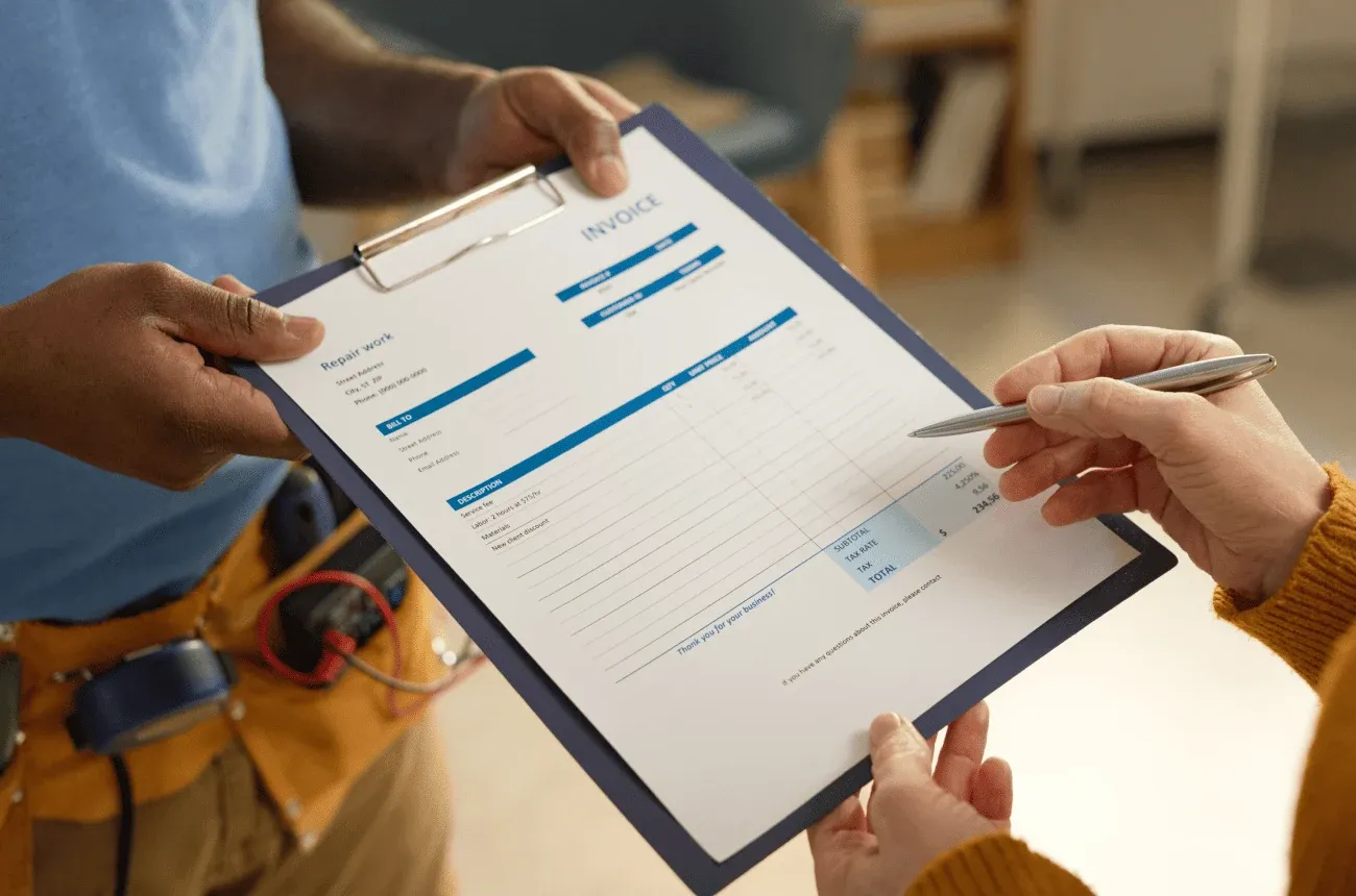 A worker in a tool belt holding a clipboard with a repair work invoice while a customer points at it with a pen