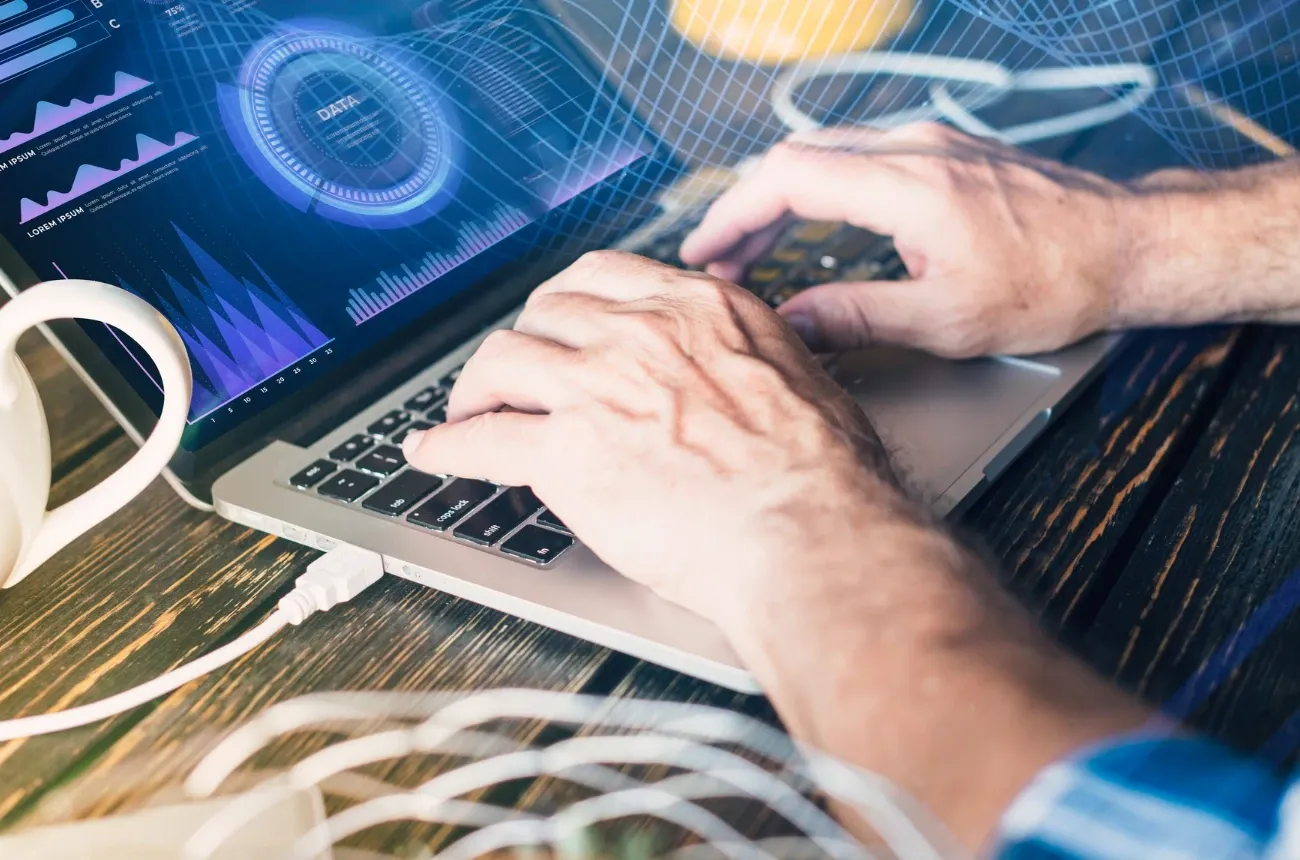 Hands typing on a laptop showing data charts, with a digital grid overlay, a white mug and a cable on a wooden desk