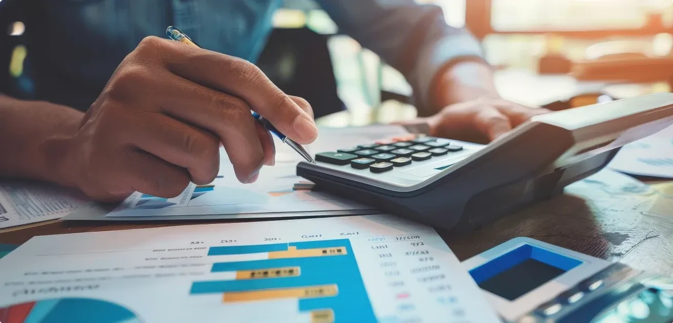 Una persona con bolígrafo revisa informes con gráficas de barras junto a una calculadora sobre un escritorio