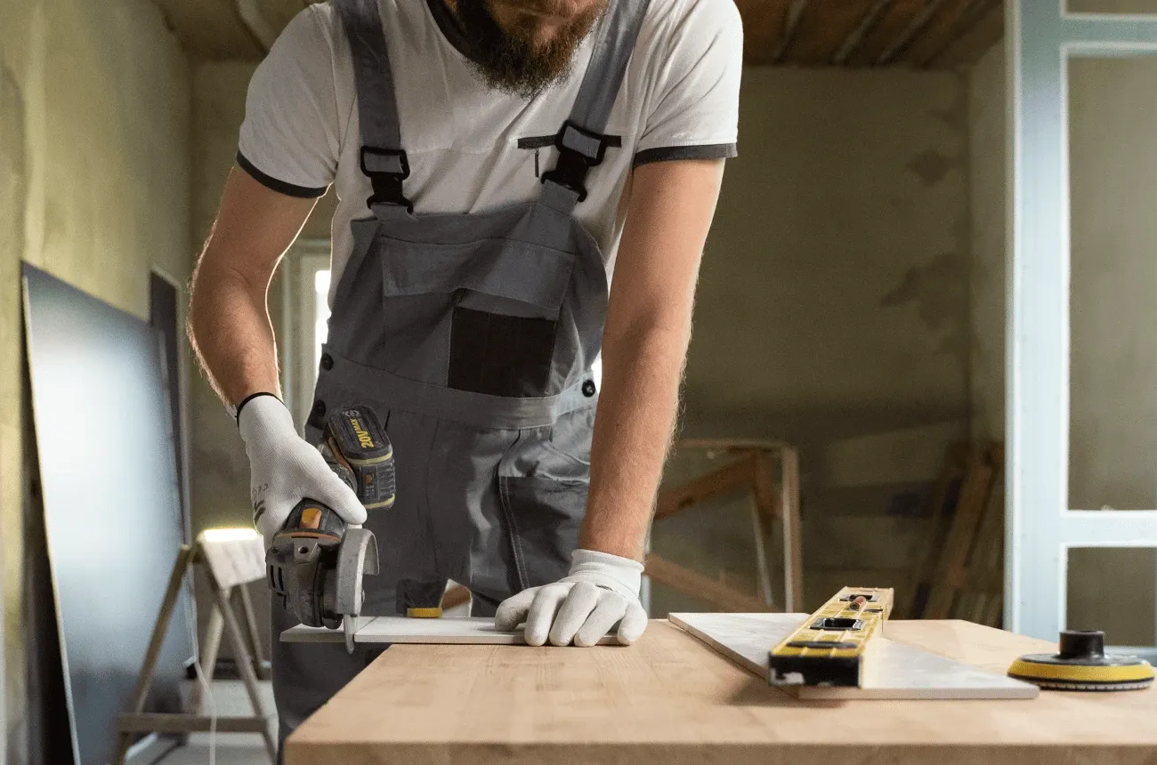 A worker in overalls and gloves cuts a tile with an angle grinder on a wooden table next to a level